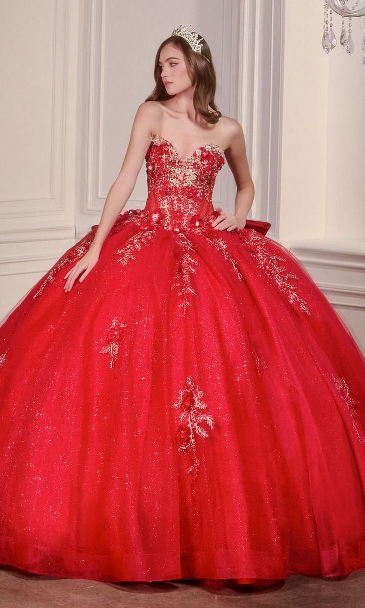 A woman in the Quinceanera Dress 15726 by Ladivine, a sparkling red ball gown with gold embellishments and glitter tulle, plus a tiara, stands in an elegant room with white paneled walls.